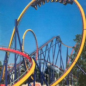 Może przedstawiać: Żółty i niebieski rollercoaster z pętlą. Kolejka górska znajduje się w parku rozrywki z ludźmi spacerującymi wokół.
