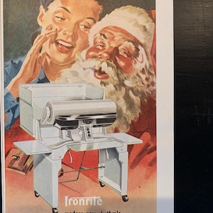 Puede incluir: Anuncio vintage con Papá Noel y una mujer, promocionando una planchadora Ironrite. La máquina es de color gris claro con una tabla de planchar incorporada. El texto dice "Tell Santa you want an Ironrite!" y "ironing means glorious freedom all year through."