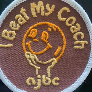 May include: Embroidered patch with the text "I beat My Coach" in yellow thread. A smiling face is in the center, also in yellow. The patch has a brown background and a white border. The letters "ajbc" are at the bottom.