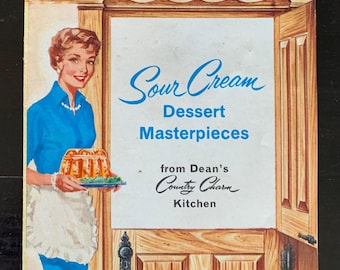 May include: A vintage cookbook cover showing a woman in a blue dress and white apron holding a dessert. The cover text reads "Sour Cream Dessert Masterpieces" from Dean's Country Charm Kitchen. The background is a wooden door frame.