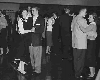 May include: A black and white photo of a group of people dancing at a school dance. The photo was taken in the 1950s. The people are dressed in formal attire and are smiling and laughing. There is a sign in the background that says "No Smoking."