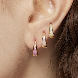 May include: Close-up of three gold hoop earrings with colourful, iridescent gemstone inlays. The earrings are in a triangular shape and are worn on an ear. The earrings have a polished finish against a blurred background.