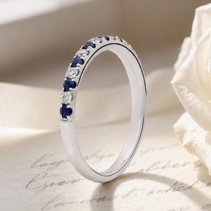 May include: A silver-toned metal ring featuring a row of alternating round-cut dark blue sapphires and clear white diamonds set along the top half of the band. The ring is positioned upright, showcasing its polished finish and gemstone arrangement.