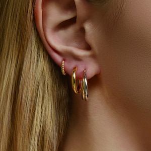 Puede incluir: Primer plano de una oreja adornada con tres pendientes diferentes. El primero es un aro pequeño de cuentas doradas. El segundo es un aro dorado más grande. El tercero es un aro plateado y dorado. Los pendientes se muestran en la oreja de una persona.