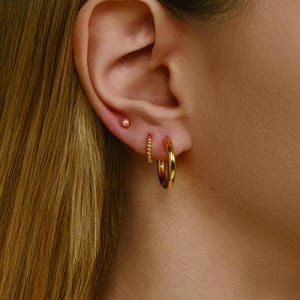 May include: Close-up of a person's ear adorned with three gold earrings. A small gold ball stud, a beaded hoop, and a larger, smooth gold hoop earring are visible. The earrings are set against the backdrop of the person's skin and hair.