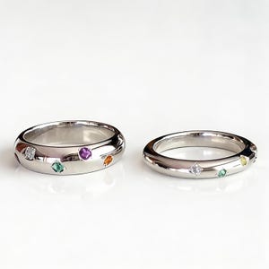 May include: Two silver rings with colorful gemstones. One ring has a wider band and features a white, green, purple, and orange gemstone. The other ring has a thinner band with a white, green, and yellow gemstone.