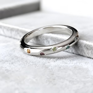 May include: A polished silver ring featuring a series of small, coloured gemstones. The gemstones include yellow, purple, green, and blue. The ring is displayed on a soft, grey surface.