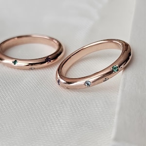 May include: Two rose gold rings with small, colorful gemstones. One ring is in focus, displaying a curved design with multiple embedded gems. The other ring is in the background, with a more traditional circular shape. The gemstones are green, purple, and blue.