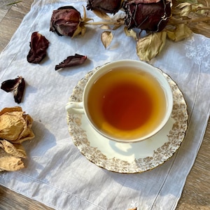 May include: A white porcelain teacup and saucer set featuring a delicate gold floral pattern along the rim, filled with amber-colored tea. The set rests on a white embroidered linen napkin surrounded by several dried dark red and beige roses.