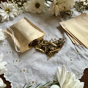 Puede incluir: Primer plano de una bolsita de té llena de hierbas secas, con algunas hierbas derramadas. La bolsita de té está sobre un paño blanco bordado, rodeada de margaritas blancas y vegetación. Varias otras bolsitas de té están apiladas cerca.