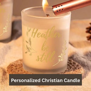 May include: A lit, frosted glass candle with gold lettering that reads "Heather be still." A lighter is held over the candle. The candle sits on a woven coaster. The text "Personalized Christian Candle" is at the bottom.