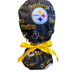 May include: A black scrub cap featuring a repeating Pittsburgh Steelers logo pattern in yellow, white, blue, and black. The fabric displays the team name and pennant graphics, and the cap is tied at the back with a bright yellow satin ribbon.