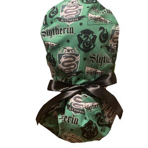 May include: Green fabric scrub cap featuring a repeating pattern of Slytherin house crests, snakes, and text. The cap is tied with a black satin bow. The text includes "Slytherin".
