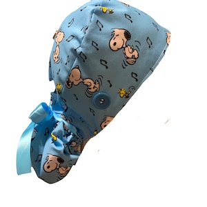 May include: A light blue scrub cap featuring a repeating pattern of Snoopy and Woodstock characters with black musical notes. The cap includes a matching light blue satin ribbon tie at the back and a single blue button on the side for mask attachment.