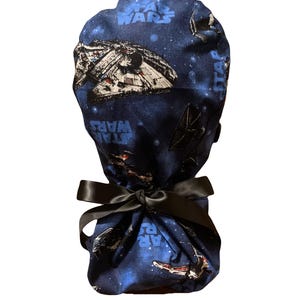 May include: A blue fabric scrub cap featuring a Star Wars print with spaceships and the text Star Wars scattered across the pattern. The cap includes a black satin ribbon tied in a bow at the back to secure the gathered fabric in place.