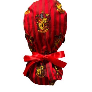 May include: Red and maroon striped scrub cap with a red satin bow. The cap features the Gryffindor crest in yellow and gold. The cap is made of fabric.