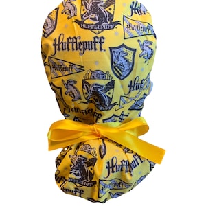 May include: A yellow Hufflepuff scrub cap with a matching yellow bow. The cap is patterned with Hufflepuff crests, badgers, and text. The design is in black and white on a yellow background. The bow is tied at the back of the cap.