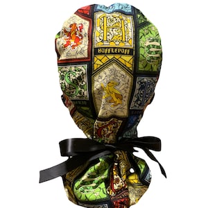 May include: A scrub cap featuring a colorful stained-glass pattern with various crests and symbols, including the text Hufflepuff. The fabric is gathered at the back and secured with a black satin ribbon tie, creating a functional head covering.