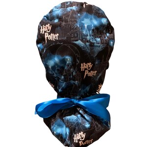 May include: A scrub cap featuring a black fabric base with a blue and white print of Hogwarts castle and the text Harry Potter. The cap includes a bright blue satin ribbon tied in a bow at the back to secure the fit around the head.