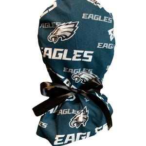 May include: A dark teal scrub cap featuring a repeating pattern of the Philadelphia Eagles logo and the word EAGLES in white text. The cap is tied at the back with a smooth black satin ribbon, creating a gathered effect at the base of the fabric.