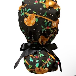 May include: A black scrub cap featuring a repeating pattern of brown sloths hanging from tree branches with green leaves and pink flowers. The cap is tied with a black satin bow.
