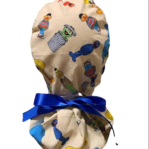Op de afbeelding: Een beige scrub cap met een kleurrijke Sesame Street-personageprint. De cap is vastgebonden met een felblauw satijnen lint. Het ontwerp bevat personages als Bert, Ernie en Oscar de Mopperkont.