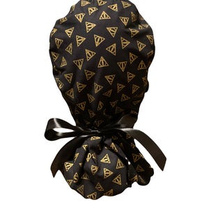 May include: A black scrub cap featuring a repeating gold pattern of the Deathly Hallows symbol. The cap has a gathered back section and is secured with a black satin ribbon tied into a bow at the nape, providing an adjustable fit for the wearer.