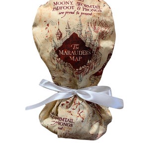 May include: A cream-colored fabric item featuring a map design with a castle illustration and the text "The Marauders Map." The item is tied with a white satin ribbon. The text "Messrs Moony, Wormtail, Padfoot & Prongs are proud to present" is also visible.