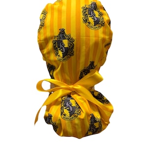 May include: A yellow and gold scrub cap with a striped pattern and the Hufflepuff crest. The cap is tied with a yellow ribbon. The Hufflepuff crest features a badger and the words "Hufflepuff".