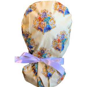 May include: A light yellow scrub cap featuring a repeating pattern of a crest with the letter "H" in the center. The crests are in blue, orange, and gold. The cap is tied with a white satin ribbon.