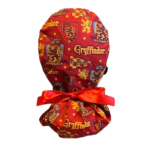May include: A red fabric scrub cap featuring a repeating pattern of gold and maroon Gryffindor crests and text. The cap is tied with a red satin bow. The cap is designed to be worn by medical professionals.