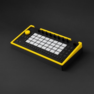 Peut inclure: Appareil de production de musique électronique noir et jaune avec une grille de boutons blancs, des boutons rotatifs et un grand bouton jaune. L'appareil rectangulaire est conçu pour la création et la performance musicale.