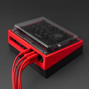 Op de afbeelding: Een rood en zwart audioapparaat met een doorzichtige plastic hoes. Het apparaat heeft meerdere knoppen en poorten, met rode kabels aangesloten. De tekst "Analog Heat MKII" is zichtbaar.