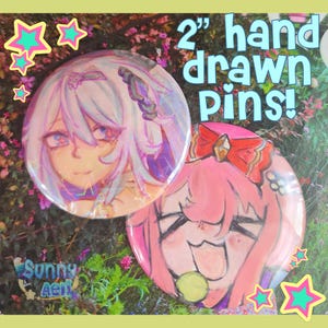 May include: Two 2-inch hand-drawn pins featuring anime-style character illustrations. One pin has a light purple and blue color scheme, while the other is pink with a red bow. The pins are set against a floral background.