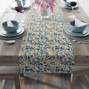 May include: A decorative table runner with a cream backdrop and a repeating pattern of navy, yellow, and red flowers and leaves. The runner is laid on a wooden table set for a meal, with glasses and tableware. A vase of pink lilies is in the centre.