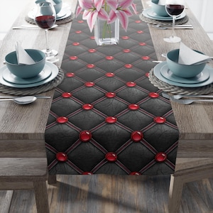 Puede incluir: Un camino de mesa negro con un patrón de diamantes y detalles en rojo. El camino de mesa presenta un diseño acolchado con adornos circulares rojos en cada intersección del patrón de diamantes. El camino de mesa está sobre una mesa de madera con cubiertos.