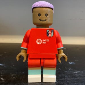 May include: A 3D-printed plastic toy figure with a tan head, lavender hair, and a red jersey. The jersey features a United Way logo and a KC crest on the chest. The figure has tan hands, red shorts, light blue socks, and white shoes with a textured finish.