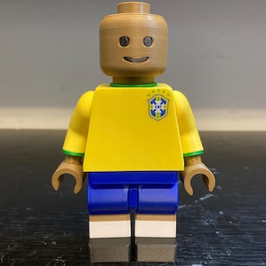 May include: A plastic toy figure resembling a building block character with a tan head, a yellow torso featuring a green and blue Brazil football crest, blue shorts, white socks, and dark grey shoes. The figure has a simple printed face with eyes and a smile.