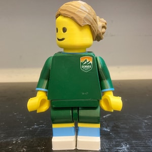 May include: A custom Lego minifigure with a yellow head and hands, wearing a green shirt and shorts with blue and white accents. The shirt features a Denver logo. The hair is light brown.