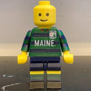 May include: A yellow plastic toy figure with a cylindrical head and a smiling face. The torso features horizontal green and navy blue stripes with the word MAINE printed in white and a Portland logo. The arms are striped, and the legs are navy blue.