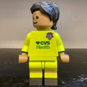 May include: A plastic toy figure with tan skin, dark hair in a ponytail, and a bright yellow soccer jersey. The jersey features a black Washington Spirit logo on the chest and green CVS Health text with a heart icon. The figure has grey shoes.