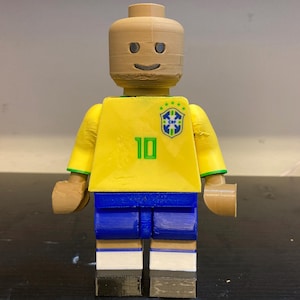 May include: A 3D-printed figure of a footballer wearing a yellow jersey with the number 10 and a Brazilian flag emblem. The figure has blue shorts, white socks, and brown skin and hair, resembling a Lego minifigure.