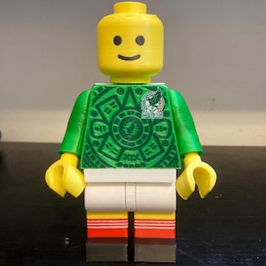 May include: A yellow plastic toy figure with a cylindrical head and a smiling face. The figure wears a green torso featuring a circular Aztec-inspired pattern and a small emblem, white shorts, and red and white striped socks with black feet.