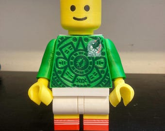 May include: A yellow plastic toy figure with a cylindrical head and a smiling face. The figure wears a green torso featuring a circular Aztec-inspired pattern and a small emblem, white shorts, and red and white striped socks with black feet.