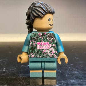 May include: A custom Lego minifigure of a person wearing a teal and floral jersey with the text "CVS Health". The figure has black hair in a ponytail, tan hands, and teal shorts. A unique, collectible item.