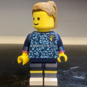May include: A plastic toy figure with a yellow head and brown hair in a ponytail. The figure wears a dark blue shirt with a light blue floral pattern, the text MERZ AESTHETICS, and a small crest. It has dark blue pants with yellow and white accents.