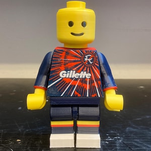 May include: A 3D printed plastic toy figure with a yellow head, yellow hands, and a torso featuring a blue and red sunburst pattern with the white text Gillette. The figure has dark blue arms, dark blue legs, and white feet with red and yellow stripes.