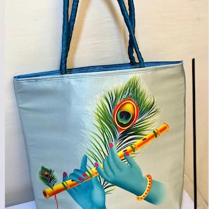May include: A light blue tote bag with teal handles, featuring a detailed illustration of blue hands playing a yellow flute, with a peacock feather. The bag measures 16 inches by 16 inches.