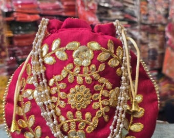May include: A red velvet pouch with gold embroidered floral designs and a beaded drawstring closure. The pouch is adorned with gold embellishments and a multi-strand pearl necklace. The background is blurred, suggesting a shop setting.