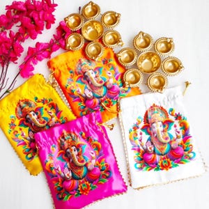 May include: An assortment of colorful drawstring bags featuring a vibrant illustration of the Hindu deity Ganesha. The bags are in yellow, orange, pink, and white, with gold-colored trim. Gold-colored oil lamps and pink flowers are also present.